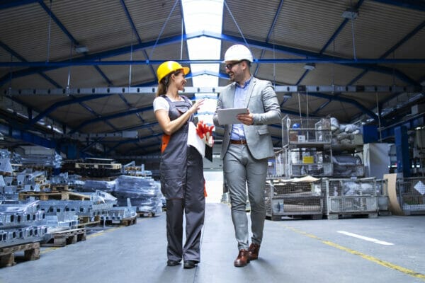 Manager supervisor and industrial worker in uniform walking in large metal factory hall and talking about increasing production.